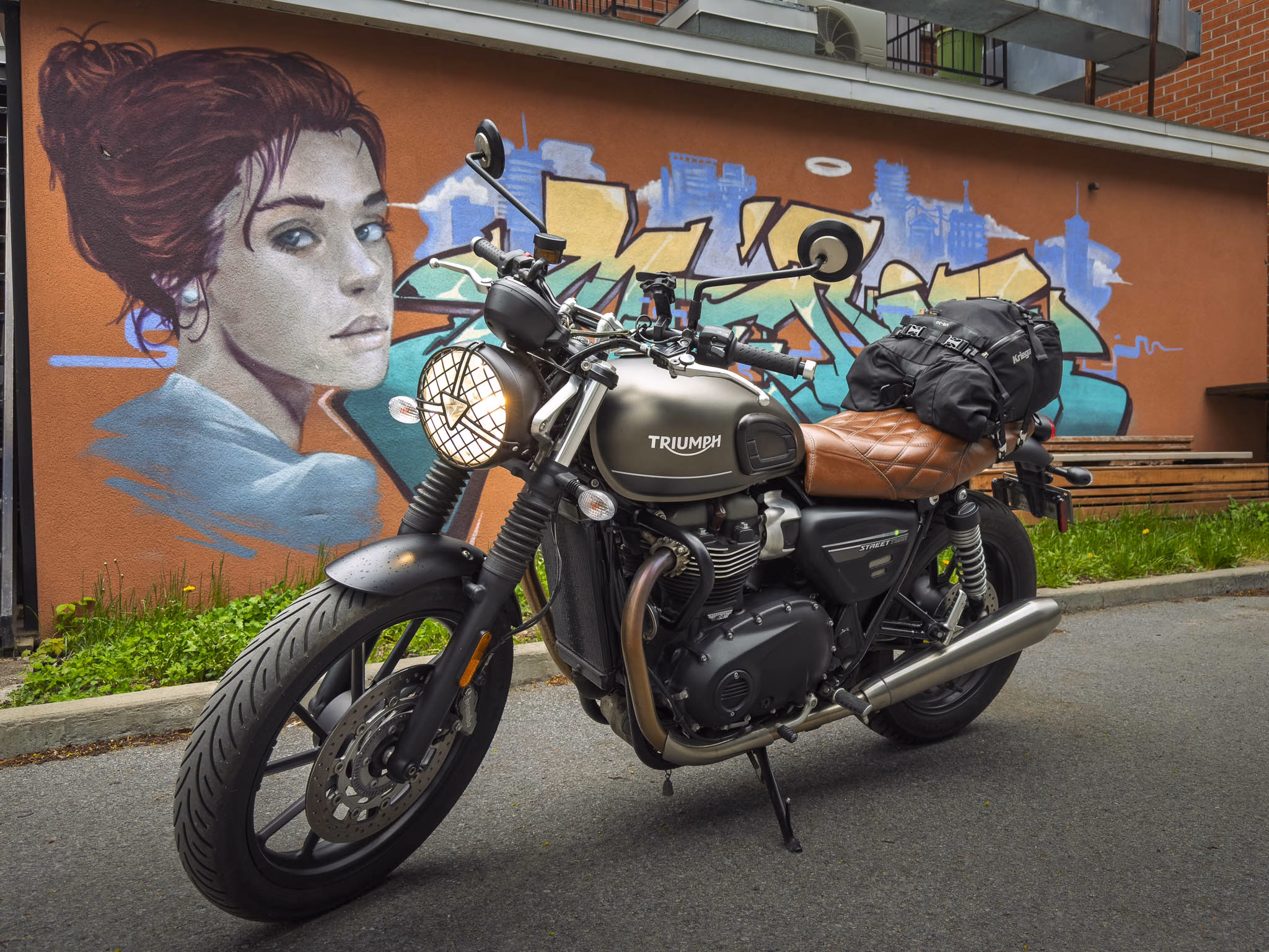 Une moto Triumph street Twin 900 devant une murale avec un visage de femme à Montréal.